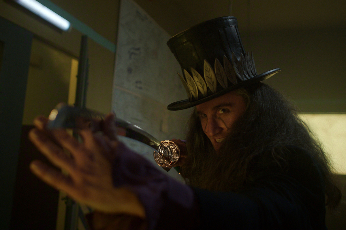A person with long hair and a tall feathered hat wields a cane or staff menacingly in a dimly lit room, with maps on the wall in the background, creating a mysterious, dramatic atmosphere.