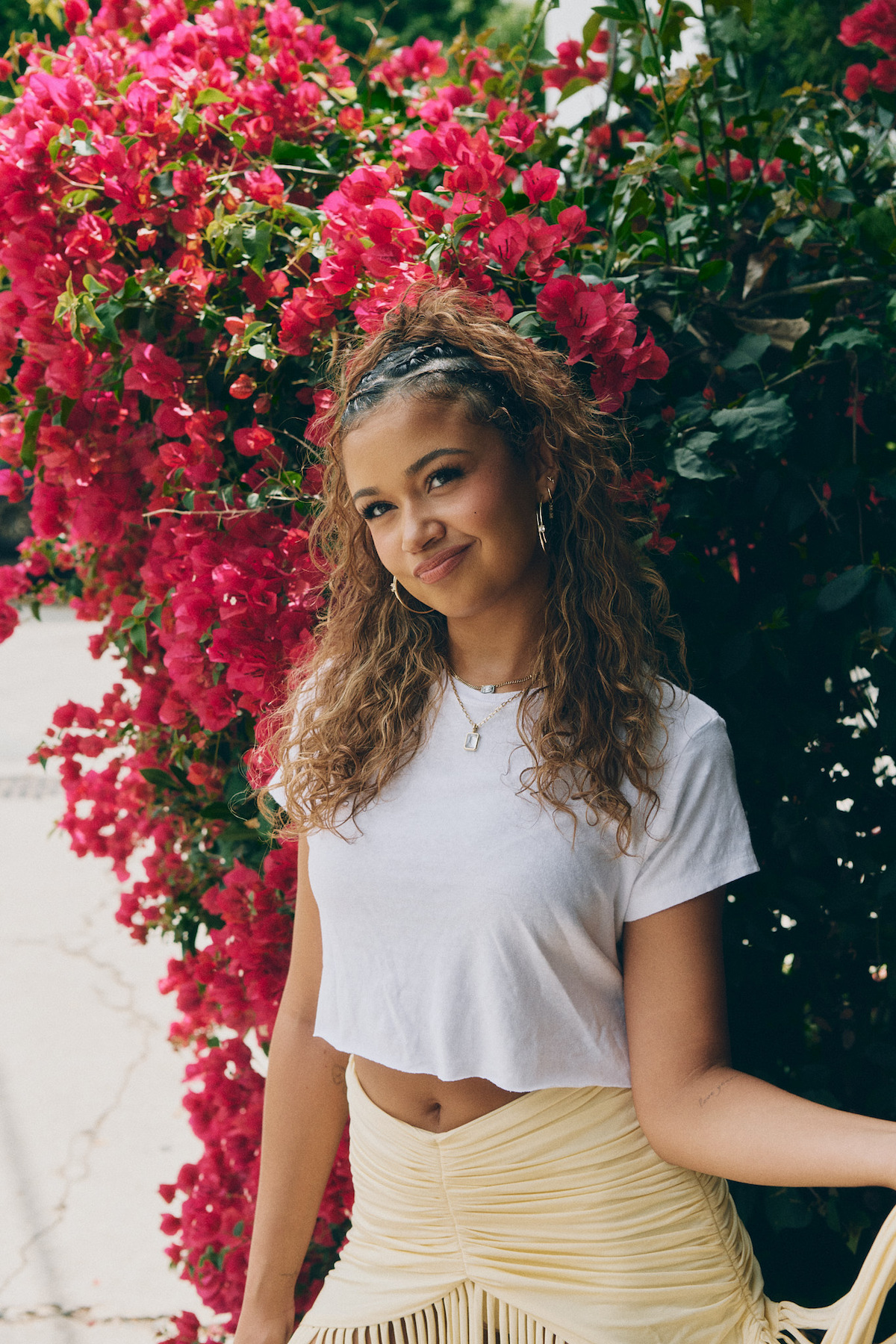 Young woman with curly hair in a white crop top and light skirt standing outdoors, smiling next to vibrant pink flowering bushes on a sunny day.