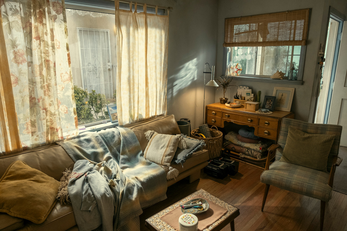 Cozy living room with sunlight through curtains, a beige sofa with blankets, a cushioned chair, a wooden desk with books and decor, a window, and a mirror reflecting the room’s warm, lived-in atmosphere.