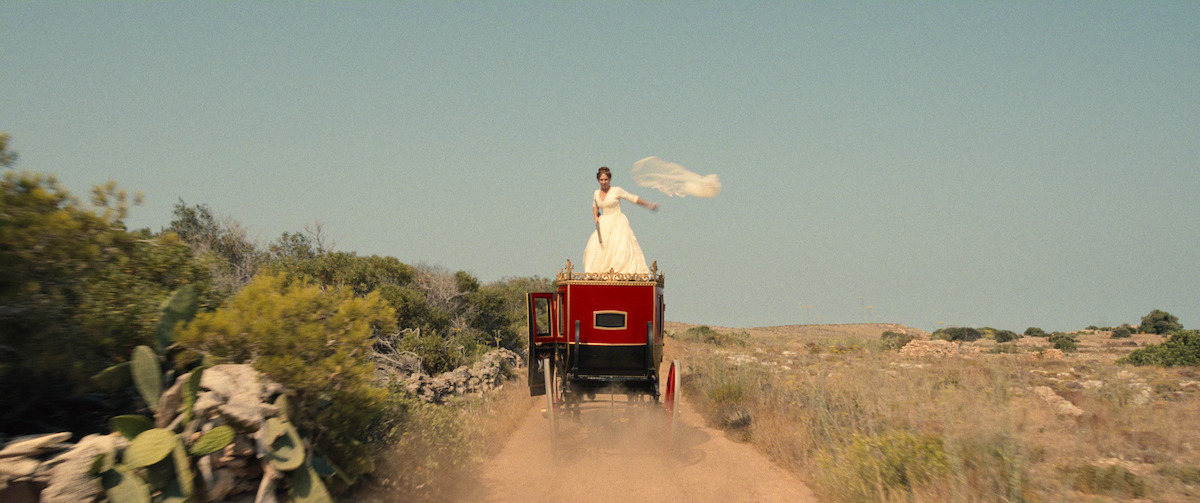 A woman in a white wedding dress stands on top of a moving red carriage on a dirt road in a rural landscape, holding a veil that flows in the wind under a clear blue sky surrounded by wild bushes and open fields.