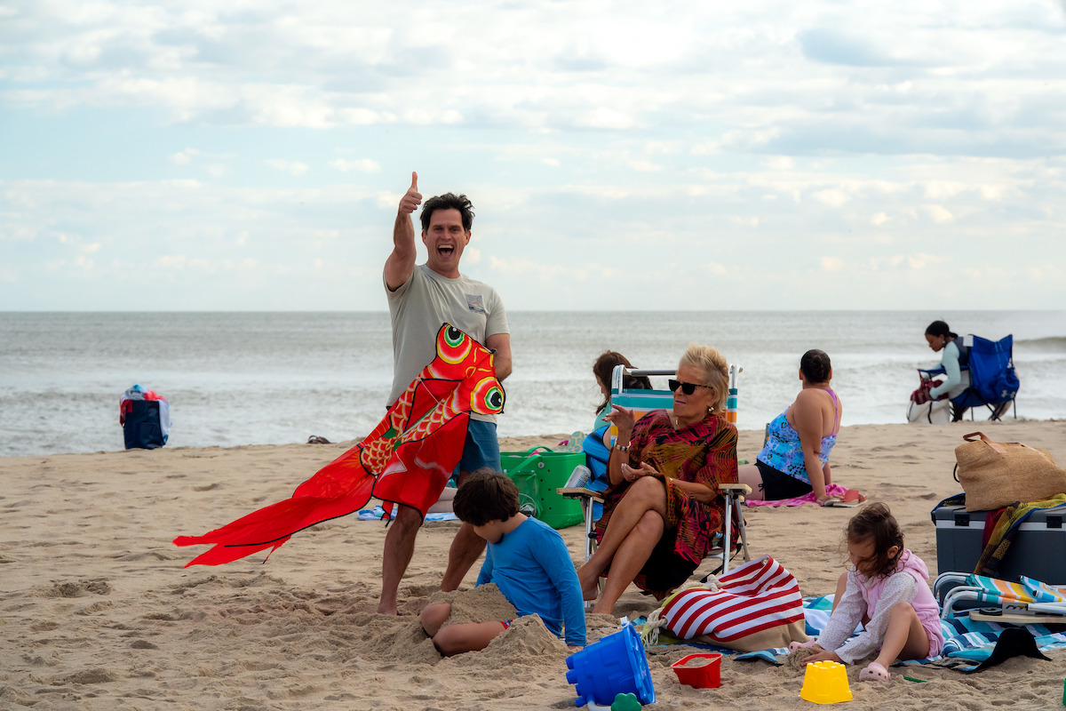 Man holding colorful kite on sandy beach, gesturing with thumb up, surrounded by seated adults and children playing, bags, and beach gear, with ocean and cloudy sky in the background. Casual family day at the beach.