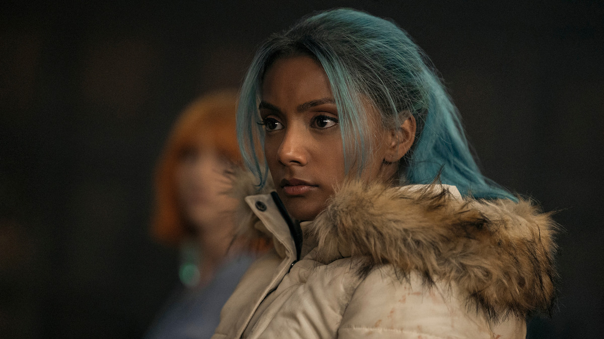 Charithra Chandran as Princess Vivi, with blue hair in a fur-trimmed winter coat stands indoors, looking serious; blurred background shows another person, suggesting a dramatic or tense moment.
