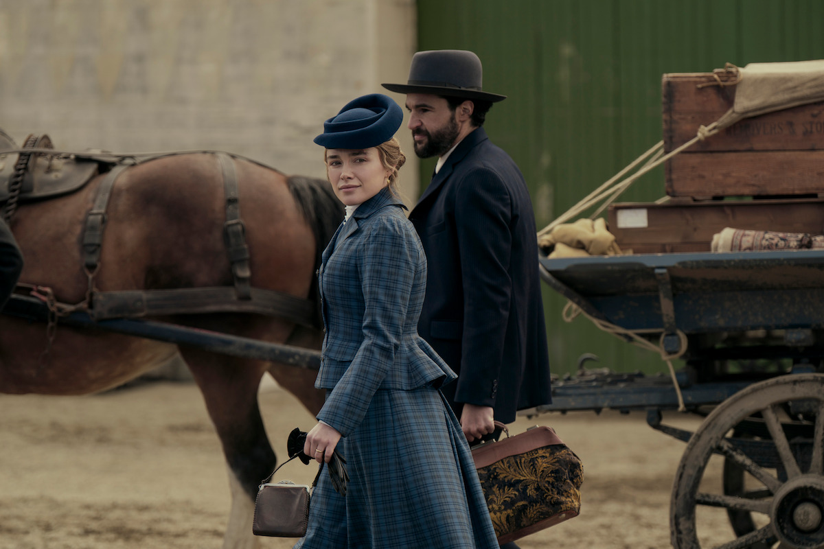 A woman and man in period clothing walk beside a horse-drawn wagon loaded with luggage and crates, set in a rustic outdoor location, evoking a historical setting.