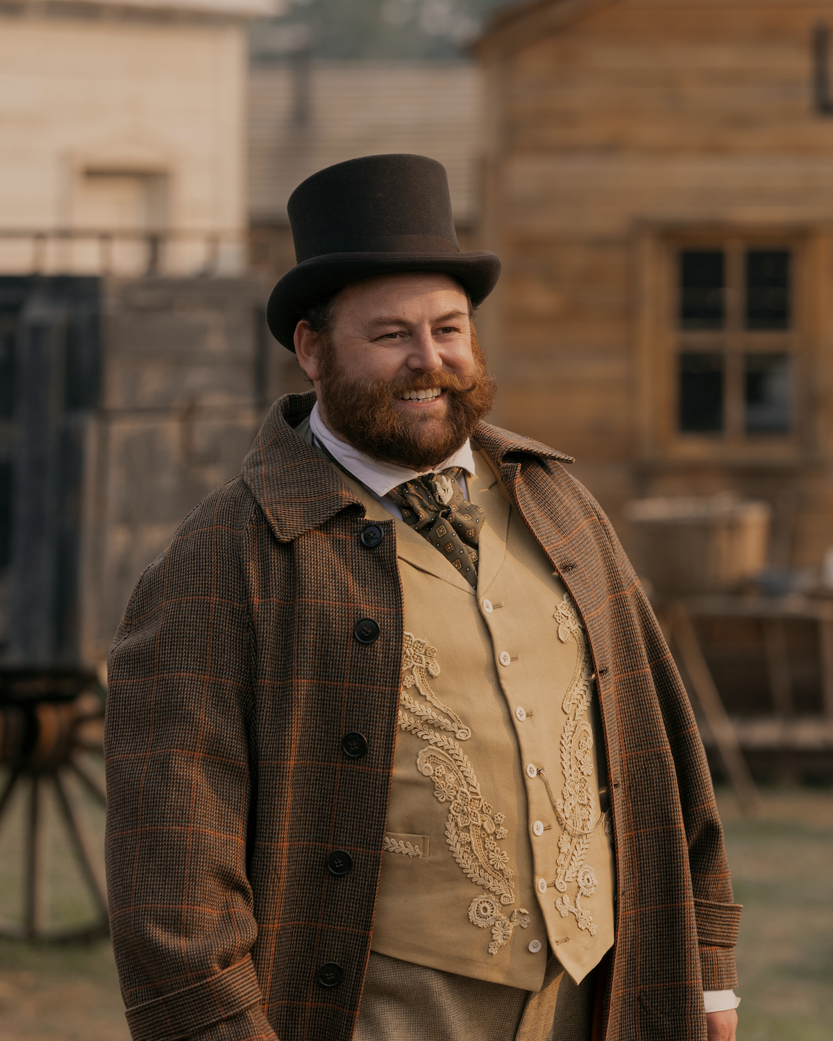 Bearded man in Victorian-era clothing with a top hat and embroidered vest smiling outdoors in a rustic, old-fashioned town setting with wooden buildings in the background.