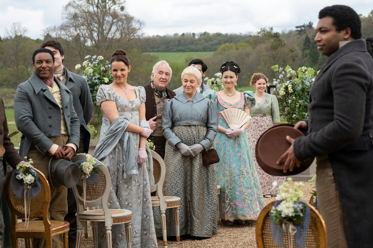 A group of people in Regency-era attire gather outdoors for a formal event, with women in elegant dresses and men in suits amid floral arrangements and countryside scenery.