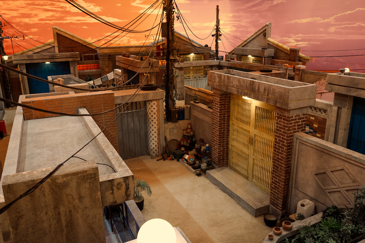 Urban alley at sunset with brick houses, power lines overhead, various pots and objects on the ground. Warm, nostalgic atmosphere suggests a quiet residential neighborhood.