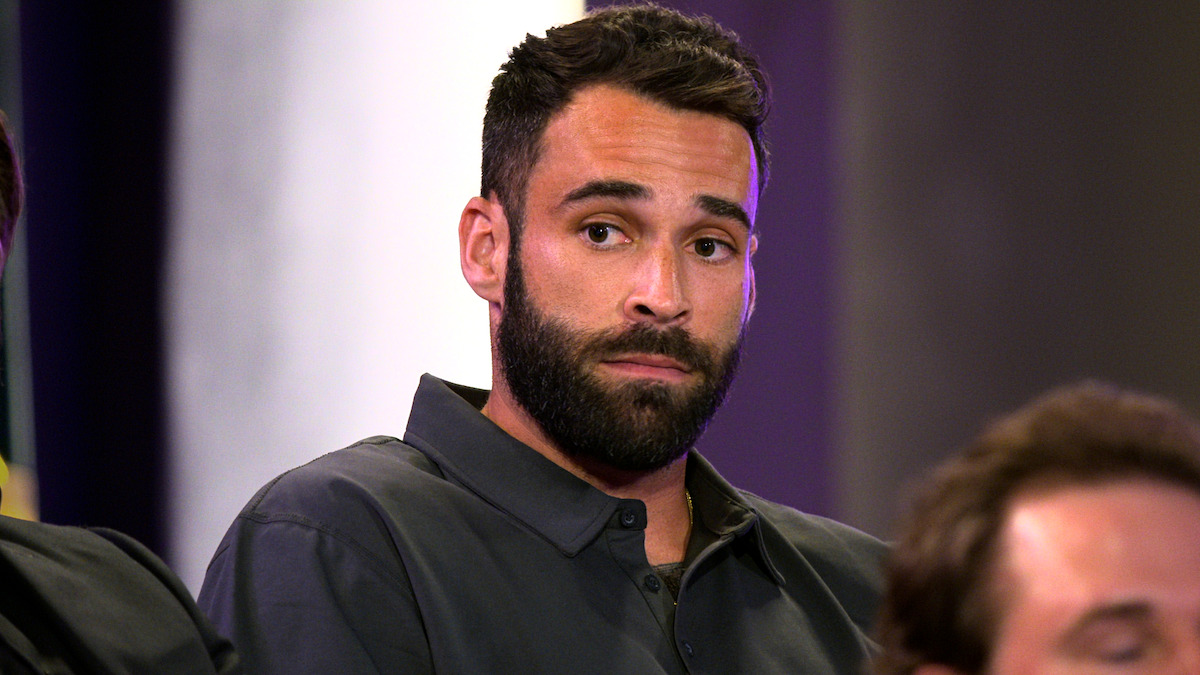 A man with a trimmed beard and dark hair, wearing a button-up shirt, sits indoors with a serious expression. The background is softly lit and blurred, with another person partially visible in the foreground.