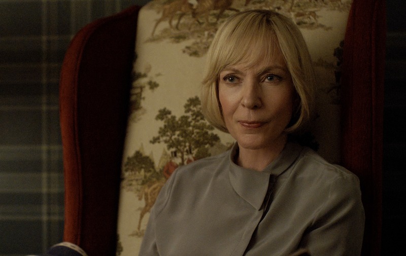 A woman with short blonde hair, wearing a light gray blouse, sits in a high-backed chair with a patterned fabric and looks thoughtfully ahead in a softly lit room.