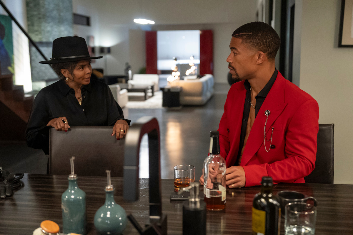 Two people sit at a dining table with drinks; a woman in a black hat and clothing faces a man in a red jacket. They are talking in a modern, well-lit living room with decorative items around.