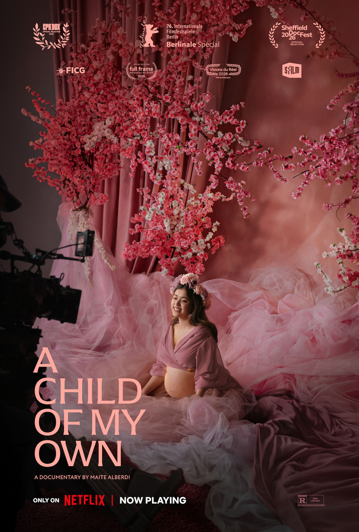 Pregnant woman in a pink dress and flower crown sits with hands on belly in a studio filled with pink tulle and cherry blossom branches, being filmed for a documentary titled "A Child of My Own," now playing on Netflix.