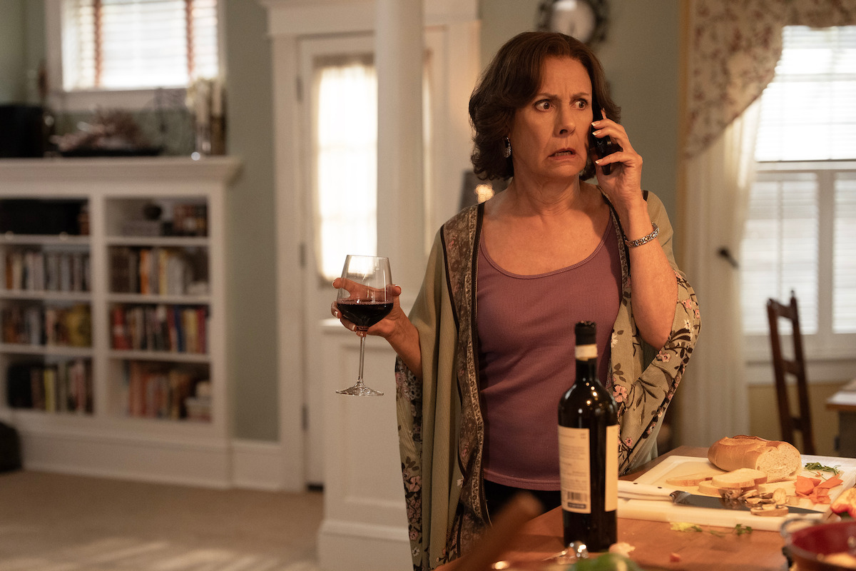 Concerned woman in a kitchen holding a glass of red wine, talking on the phone, with food and a wine bottle on the counter, shelves and windows in a warmly lit home environment.