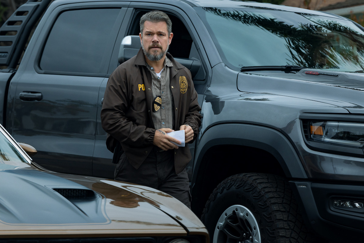 A police officer stands between two vehicles, holding papers, wearing a badge and jacket, in an outdoor urban setting with palm trees reflecting on the truck windows.