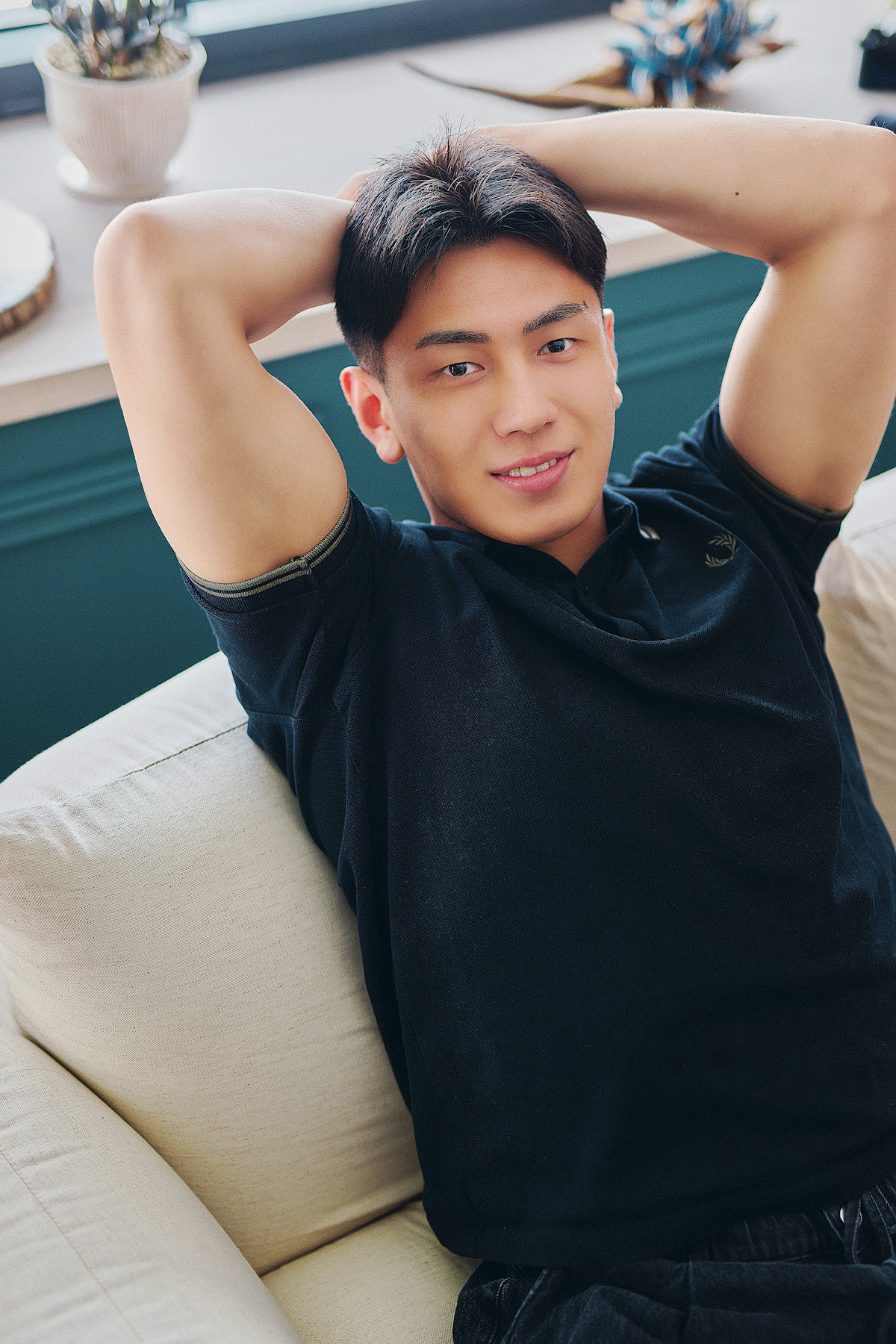 Young man relaxing on a light sofa indoors with hands behind his head, casual and comfortable atmosphere, natural light from window, calm and upbeat mood.