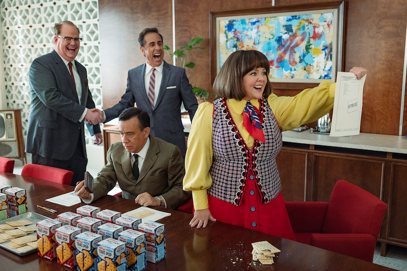 Jim Gaffigan, Jerry Seinfeld, Fred Armisen, and Melissa McCarthy smiling in a corporate office setting. 