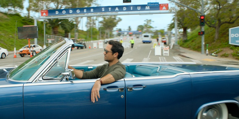 Man driving a blue convertible near Dodger Stadium entrance on a sunny day, relaxed atmosphere, street scene with greenery, traffic lights, and stadium sign in the background.