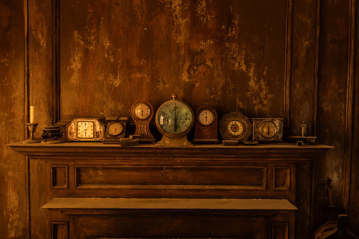 An antique mantel with multiple old clocks and a candle arranged on a rustic wooden shelf against a textured brown wall, creating a vintage, nostalgic atmosphere.