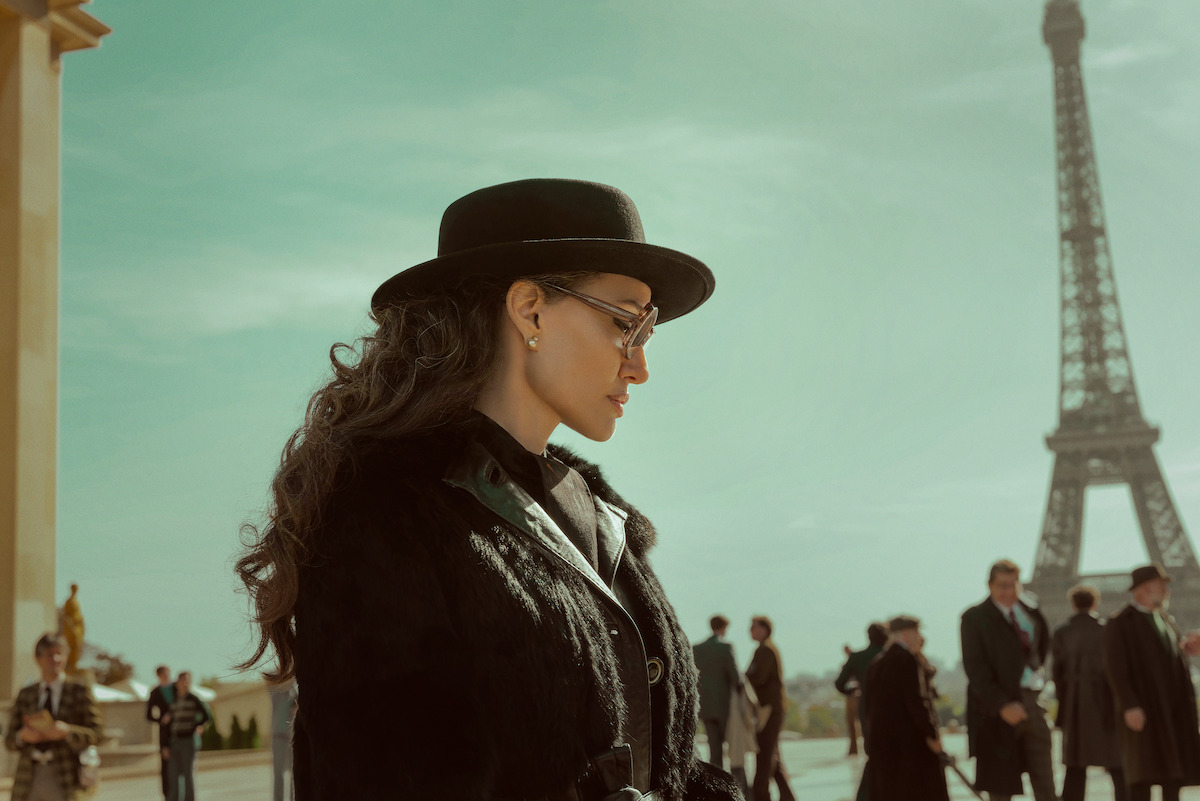 Angelina Jolie as Maria Callas wearing a hat, standing in front of the Eiffel Tower in Paris.
