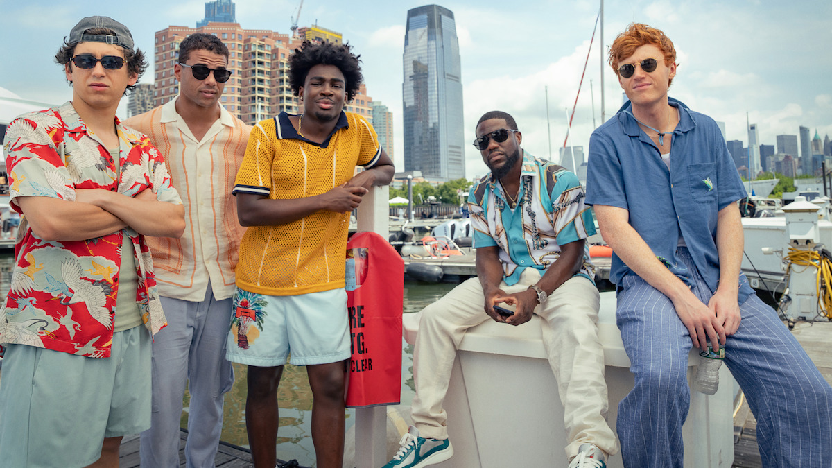 Five young men in casual summer clothes pose at a marina with sailboats, city buildings, and a skyscraper in the background, suggesting a relaxed, urban waterfront setting.