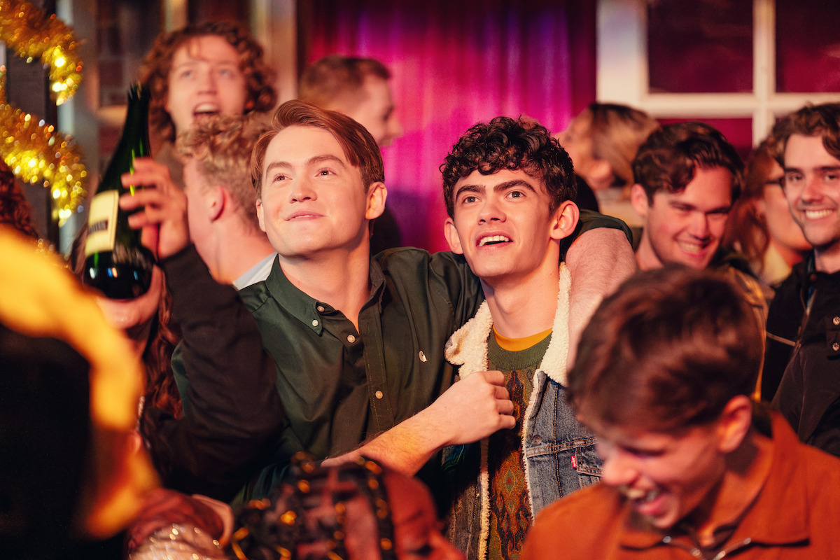 Two young men smiling and embracing at a lively indoor party, surrounded by friends celebrating in a warmly lit room with festive decorations and vibrant energy.