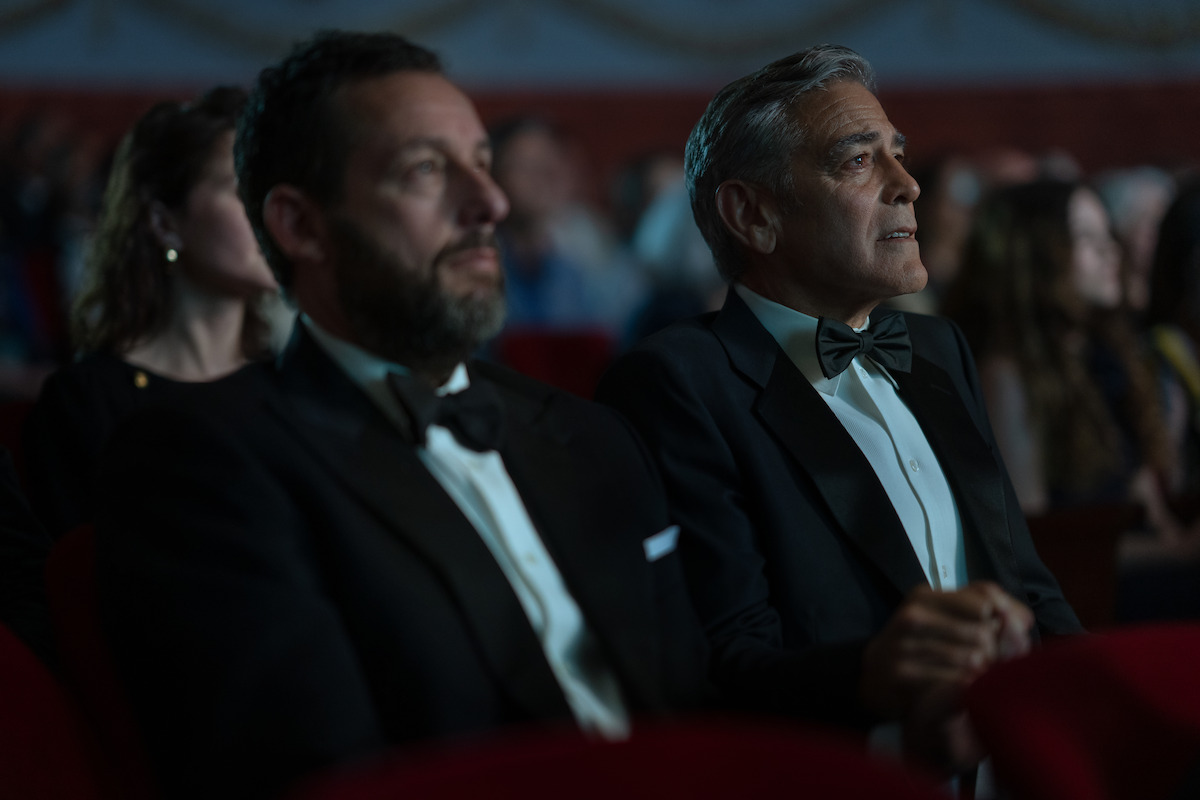 Two men in tuxedos sit in a dark theater audience, watching intently. The atmosphere is formal and focused, with other people blurred in the background, suggesting a special event or ceremony.