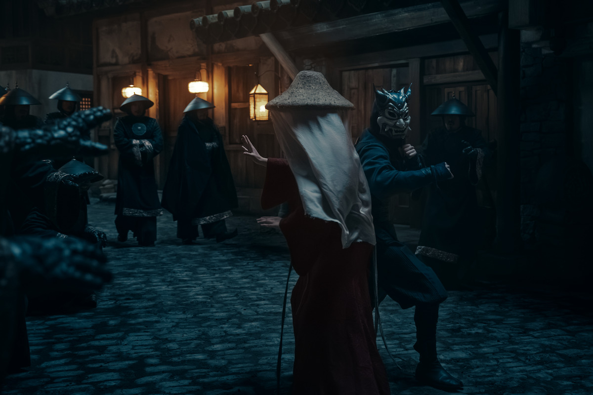 Kiawentiio as Katara and Dallas Liu as Zuko in a dimly lit cobblestone street, with lanterns illuminating wooden buildings. One wears a straw hat and cloak, another a dragon mask, surrounded by robed figures ready to fight.
