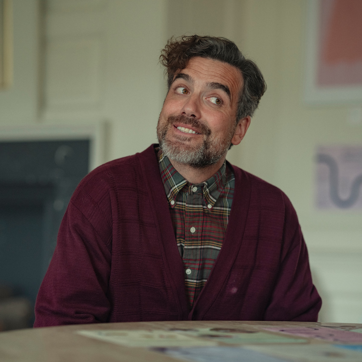 Man with gray hair and beard wearing a burgundy cardigan and plaid shirt sits at a table indoors, smiling, with soft lighting and blurred background showing wall art and neutral decor.