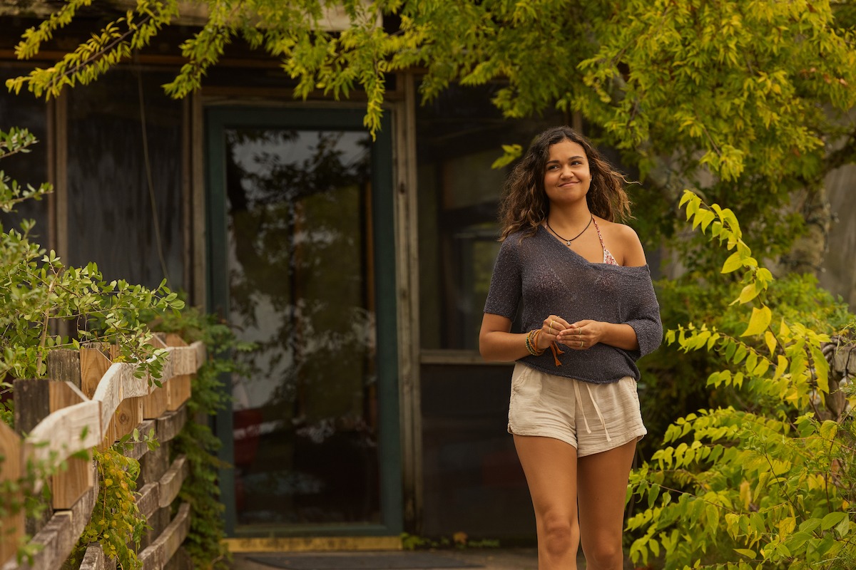 A woman stands outside a rustic building surrounded by lush green plants and wooden railings, smiling and casually dressed in a navy top and light shorts, enjoying a relaxed outdoor setting.