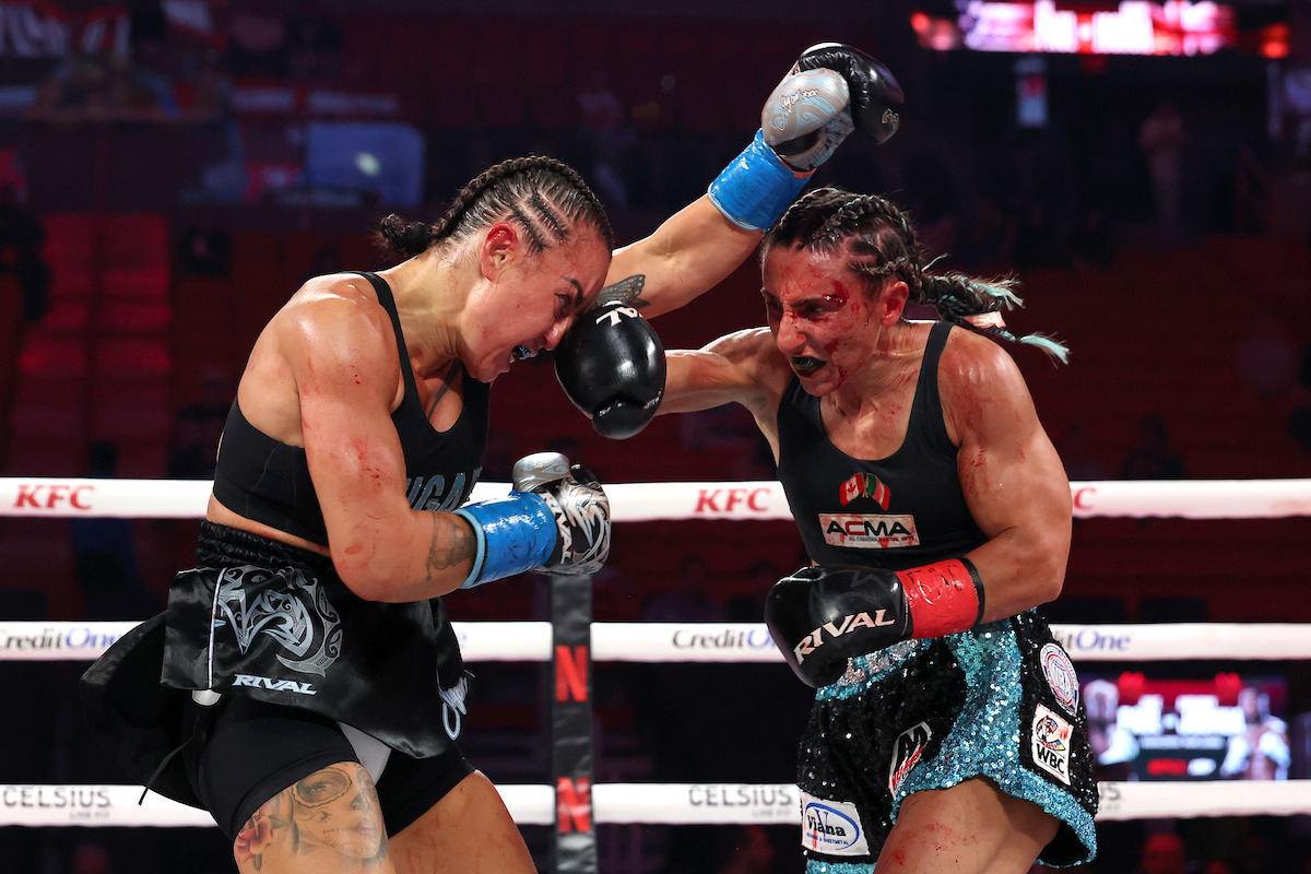 Amanda Galle and Cherneka Johnson fight intensely in a brightly lit boxing ring, one throwing a punch as both show signs of injury, sweat, and focus. The arena is filled with a dimly lit audience and branded banners.