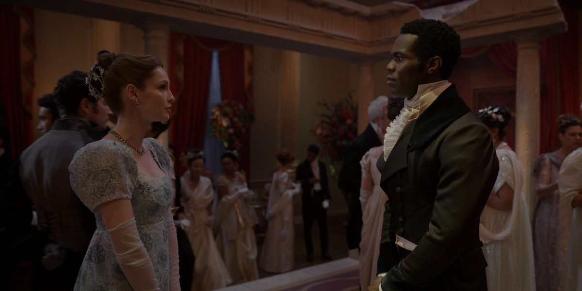A woman in a light blue gown and a man in formal attire stand facing each other at an elegant ballroom, surrounded by guests in period costumes with luxurious decor and floral arrangements in the background.