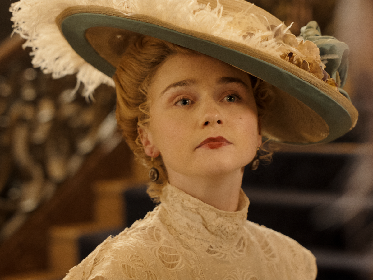Woman in vintage Edwardian attire and large feathered hat stands indoors near ornate staircase, looking serious and composed, with soft lighting highlighting her intricate lace dress.