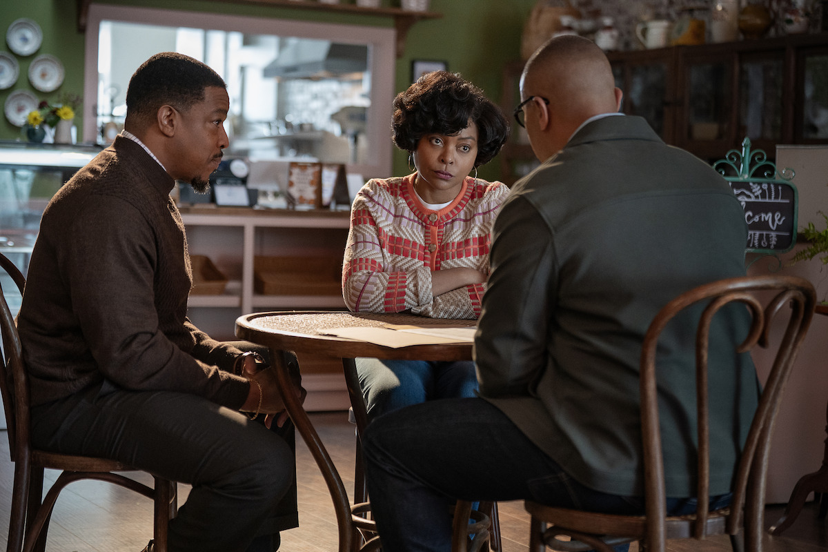 Three people sit around a small round table in a cozy cafe, engaged in a serious conversation. The setting is warmly lit, with shelves and dishes visible in the background, creating a homey atmosphere.