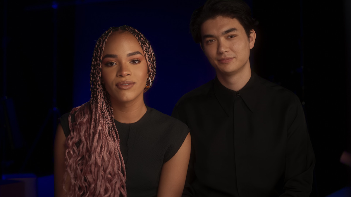 Two people sitting close together in a dimly lit studio with a dark background, both facing the camera and smiling gently, one with long pink braids and the other with short dark hair, wearing black clothing.