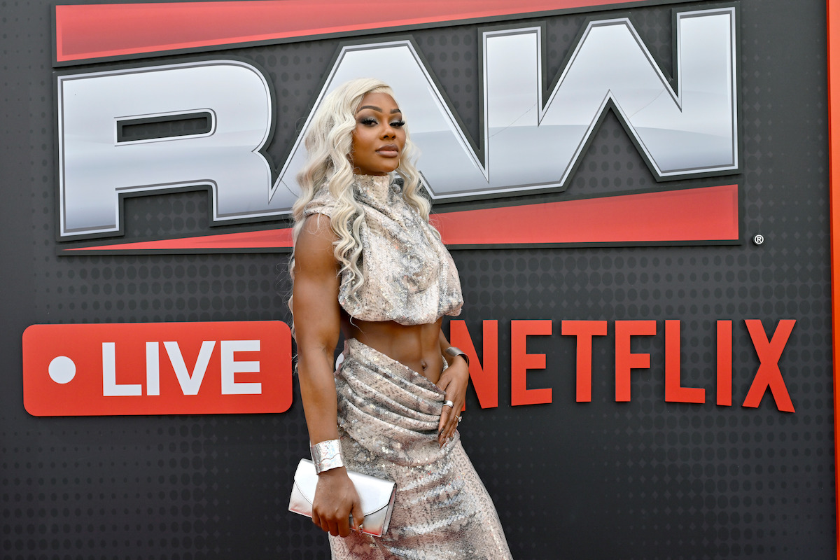 Woman with long blonde hair in a glittering silver outfit poses in front of a RAW Live on Netflix event backdrop, holding a clutch on a red carpet.
