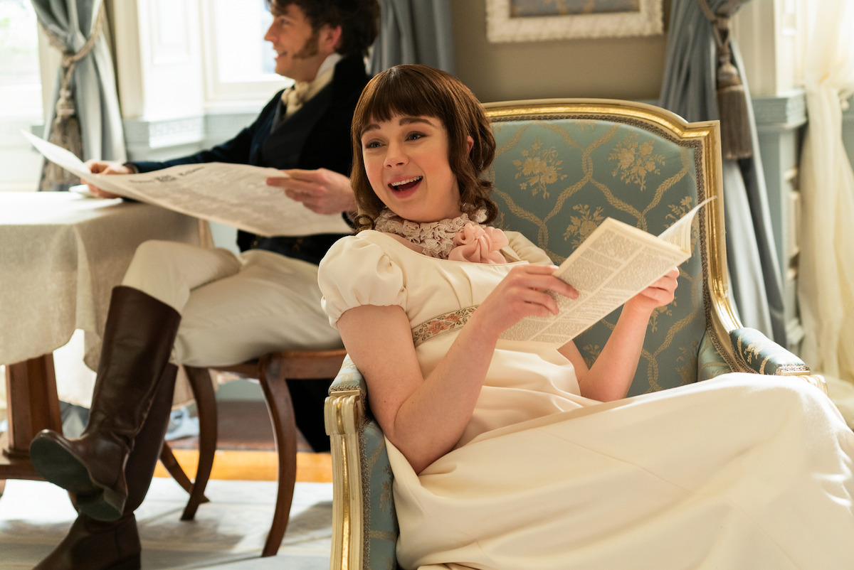 Eloise Bridgerton reclines in formal clothing on a decorative chair. She holds a piece of paper and laughs while looking off into the distance.