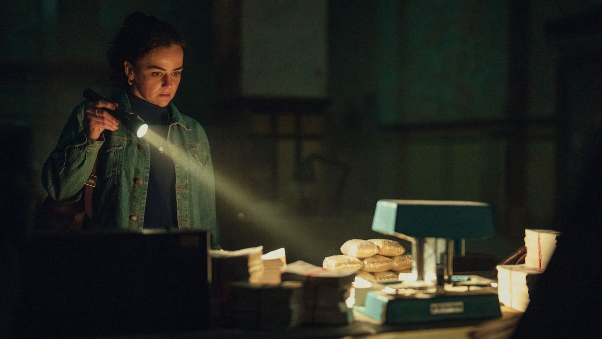 A woman in a dimly lit room shines a flashlight on a table with stacks of money, small packages, and a blue machine, suggesting investigation or discovery in a suspicious setting.