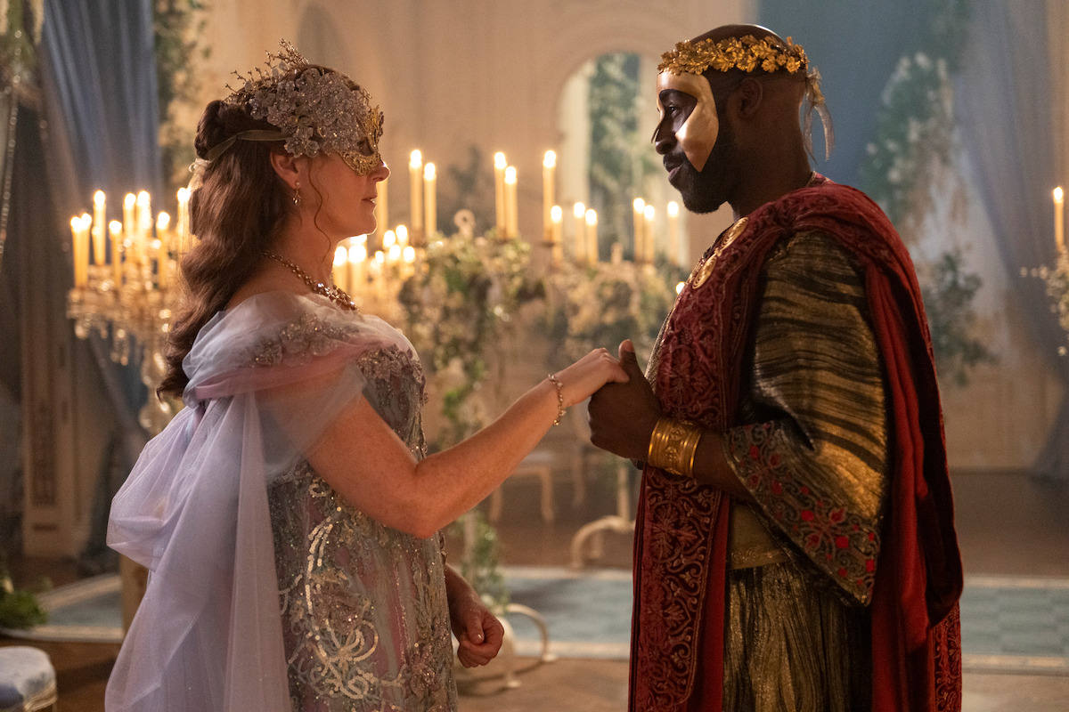 Two people in elaborate masquerade costumes hold hands in a candlelit, ornate room with draped curtains and floral decor, creating a regal and romantic atmosphere.
