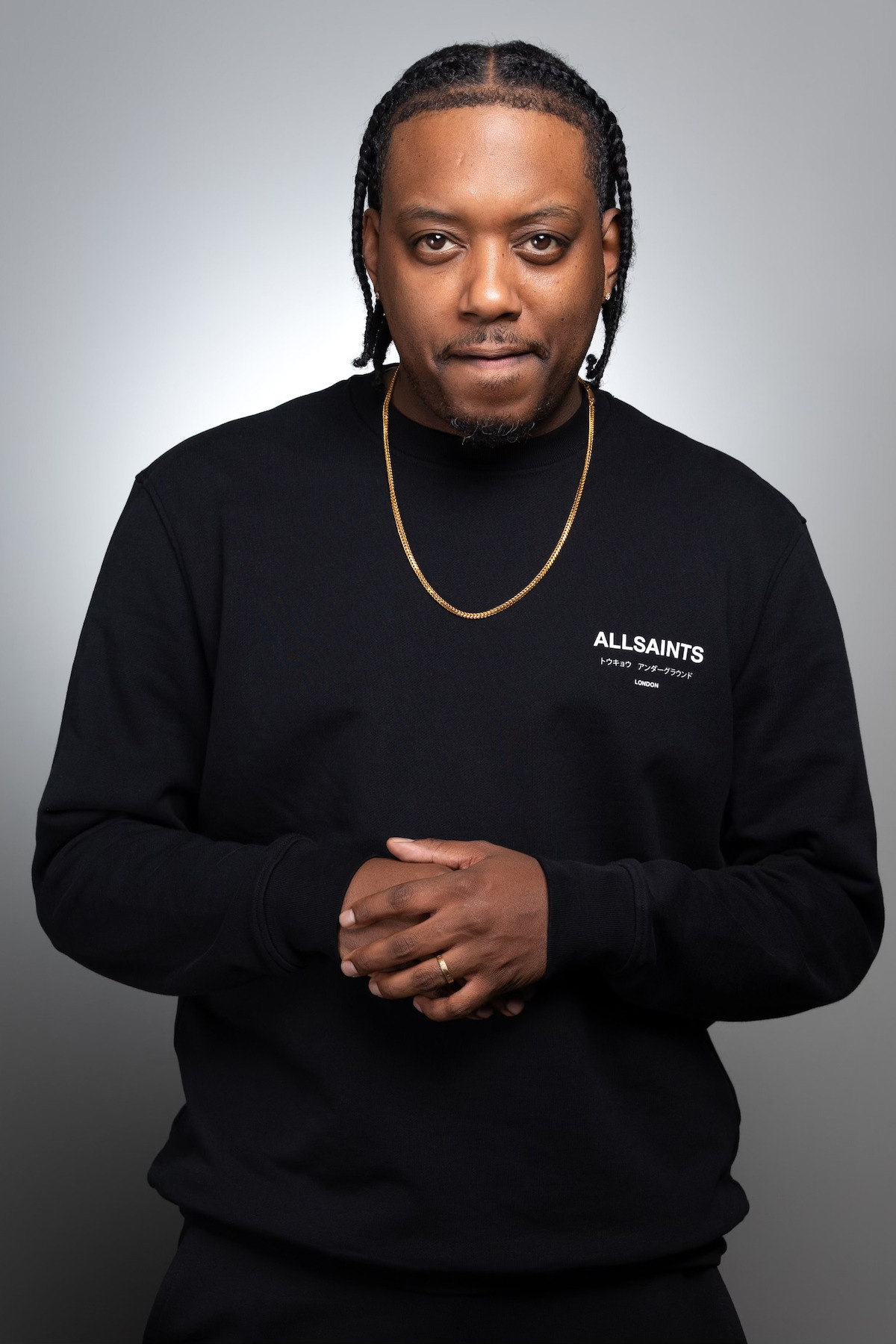 Man with braided hair wearing a black AllSaints sweatshirt and gold chain, standing against a neutral gray studio background, looking at the camera with hands clasped.