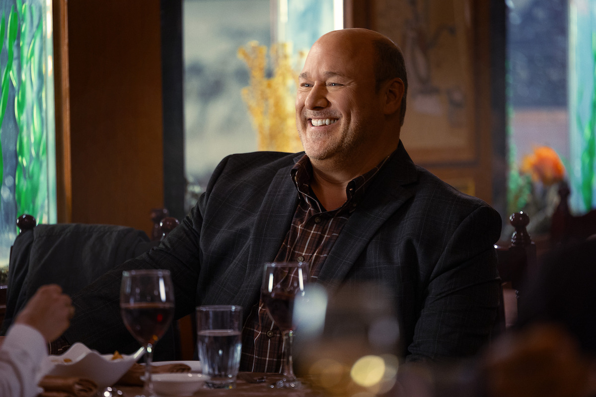 Smiling man in a dark blazer sitting at a restaurant table with wine and water glasses, colorful decor and soft lighting in the background, appearing to enjoy a social meal.