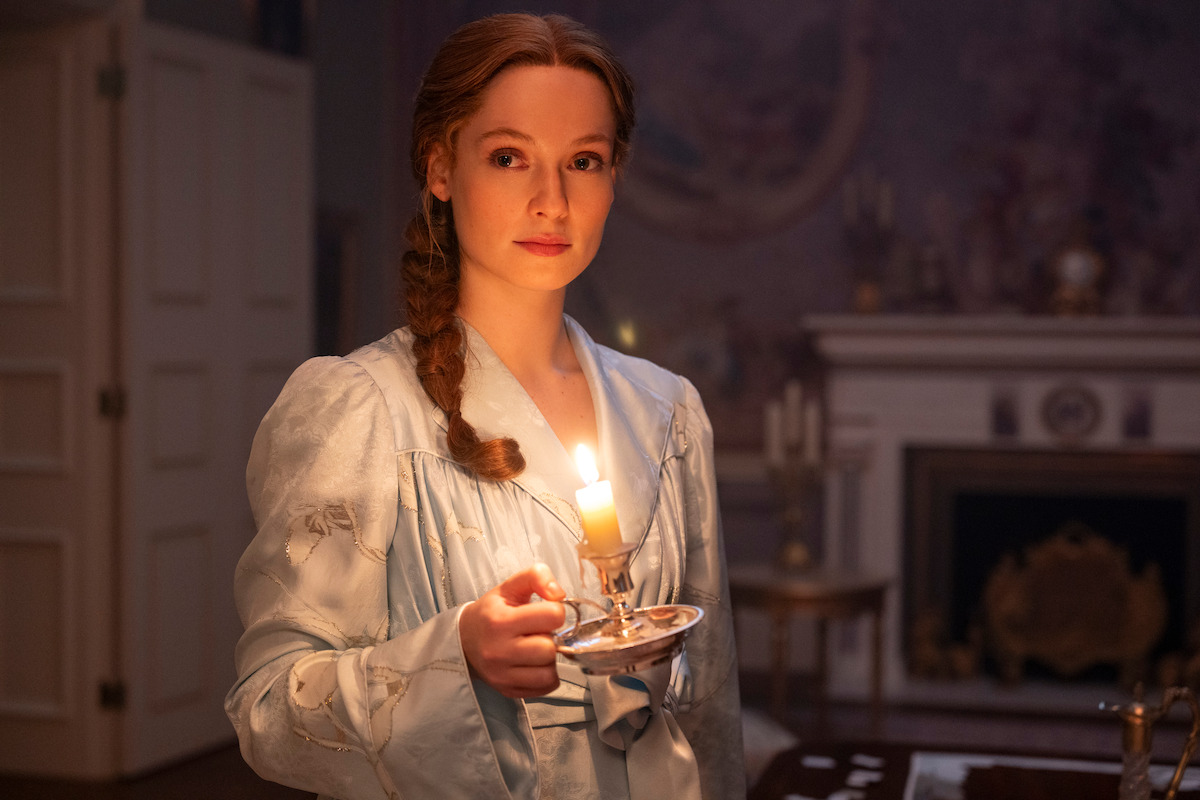 Woman in vintage clothing holding a lit candle in a dimly lit, elegant room with a fireplace and ornate decorations, evoking a historical or period setting.