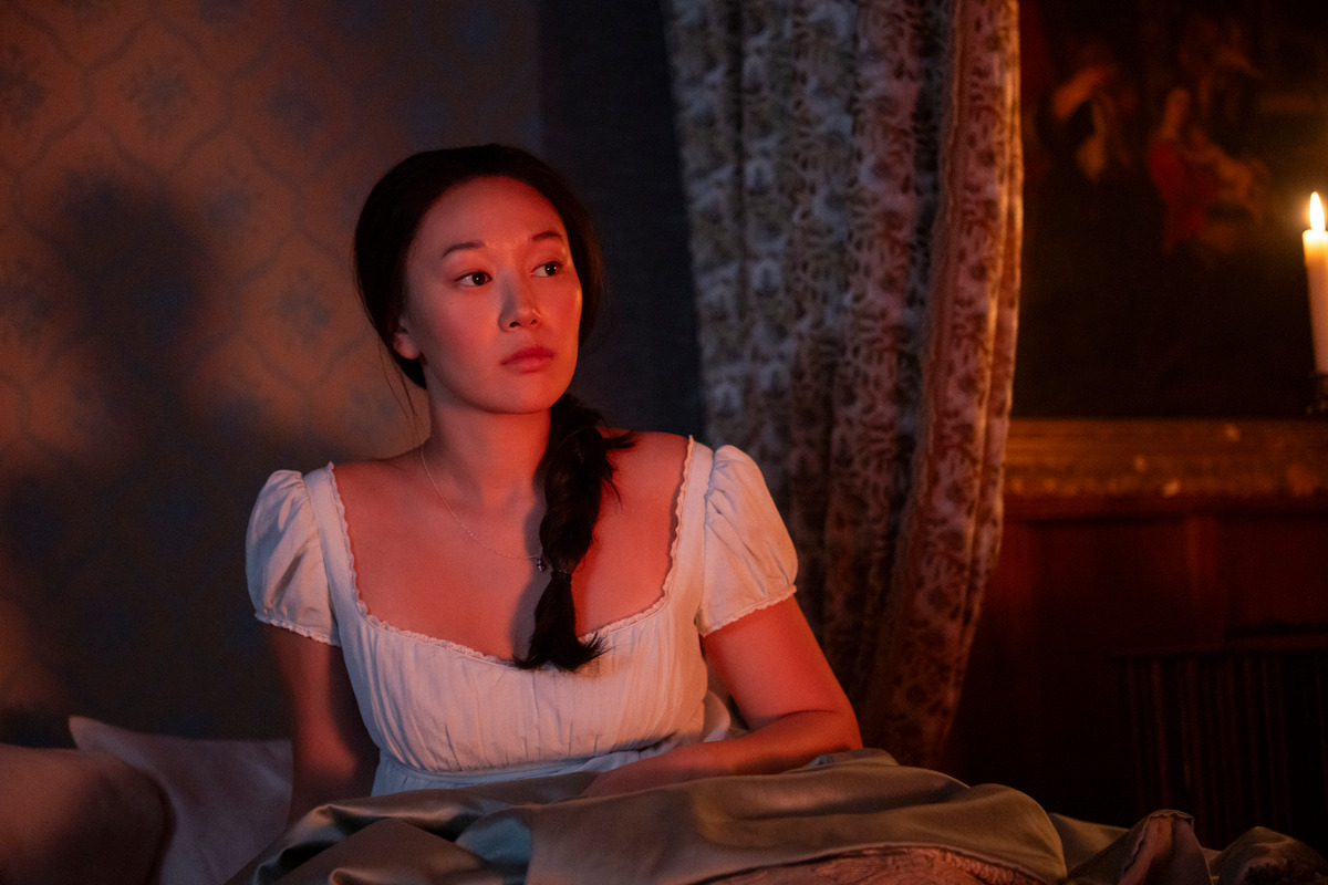 A woman in period clothing sits up in bed at night, lit by candlelight, with a thoughtful expression in a dimly lit, vintage-decorated room with curtains and a painting in the background.