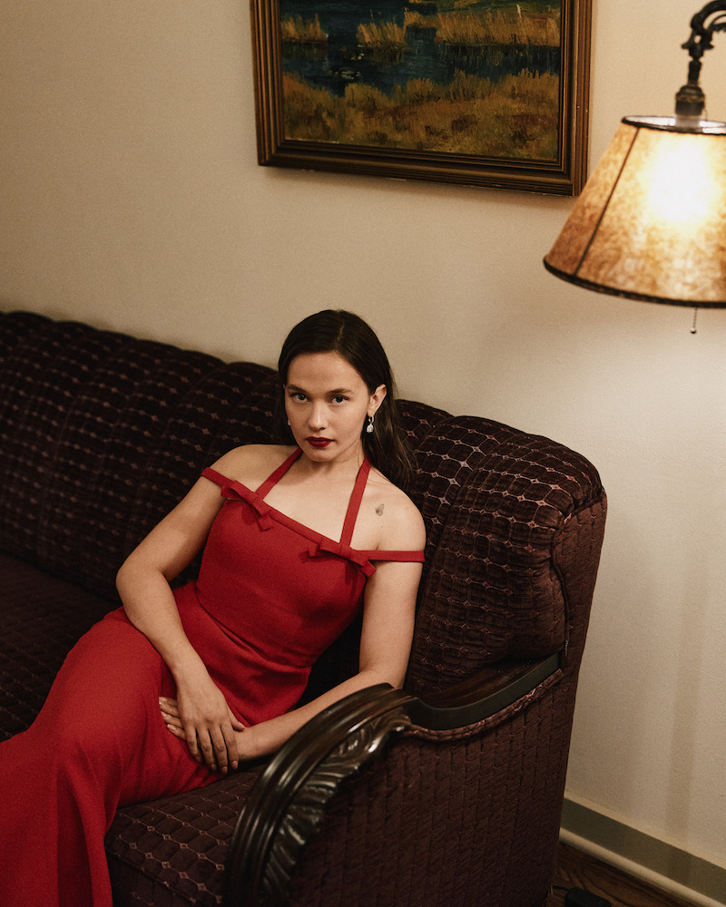 Woman in a red dress sitting on a dark patterned sofa in a warmly lit living room, with a painting and lamp on the wall behind her.