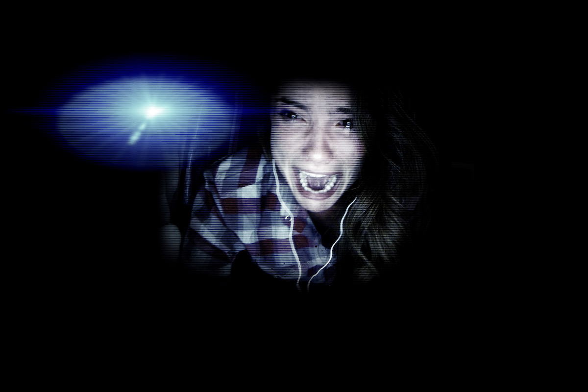 A terrified woman in a plaid shirt screams in a dark room, illuminated by bright blue-white lens flare, suggesting fear and suspense in a dramatic setting.