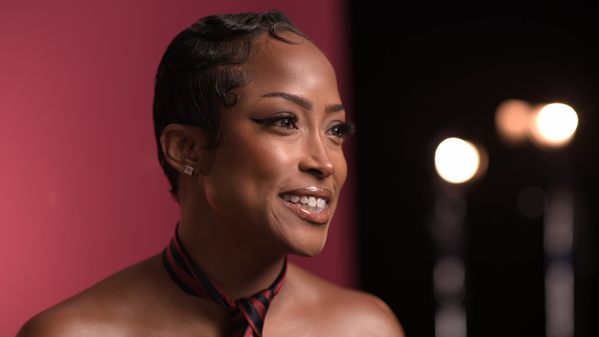 Smiling woman with short hair and a striped necktie poses in a studio with a pink and black background and blurred stage lights.