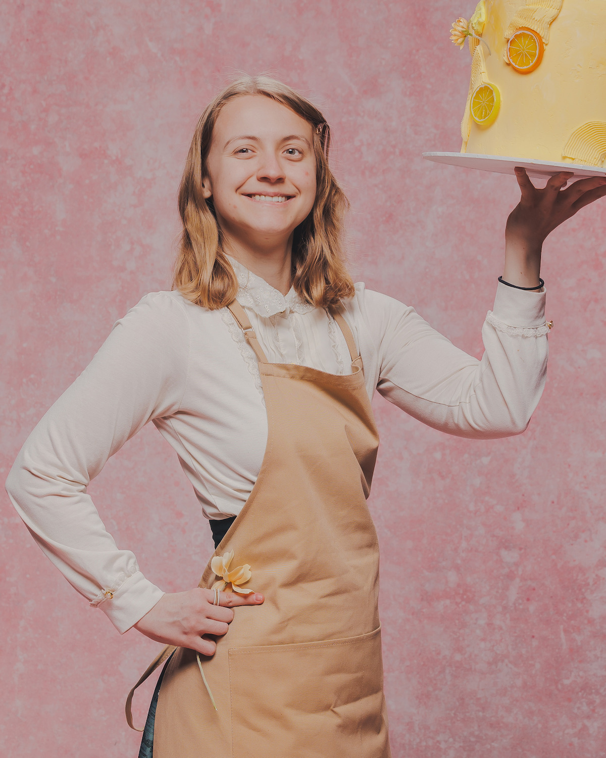 Smiling person in a tan apron holds a lemon-decorated cake on one hand, standing against a textured pink background, suggesting a cheerful bakery or baking environment.