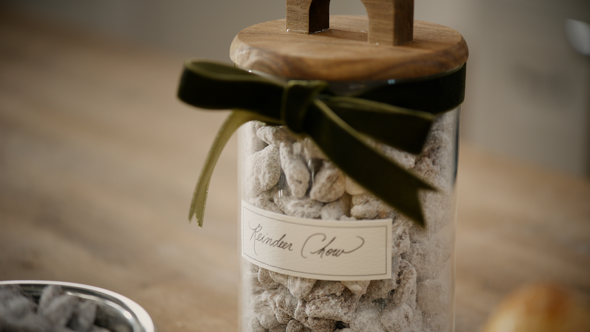 A glass jar labeled "Reindeer Chow" filled with powdered snack mix, topped with a wooden lid and green velvet ribbon, sits on a wooden surface in a cozy, softly lit kitchen setting.
