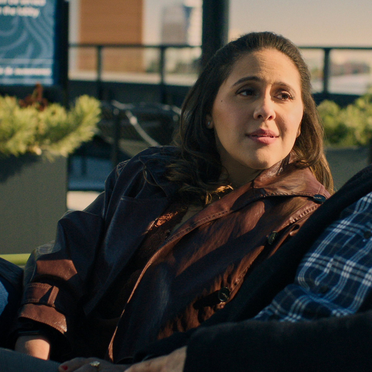 Woman in a brown jacket sits outdoors on a patio or rooftop, conversing with someone wearing a plaid shirt. The background is urban with plants and railings, suggesting a relaxed, social setting in daylight.