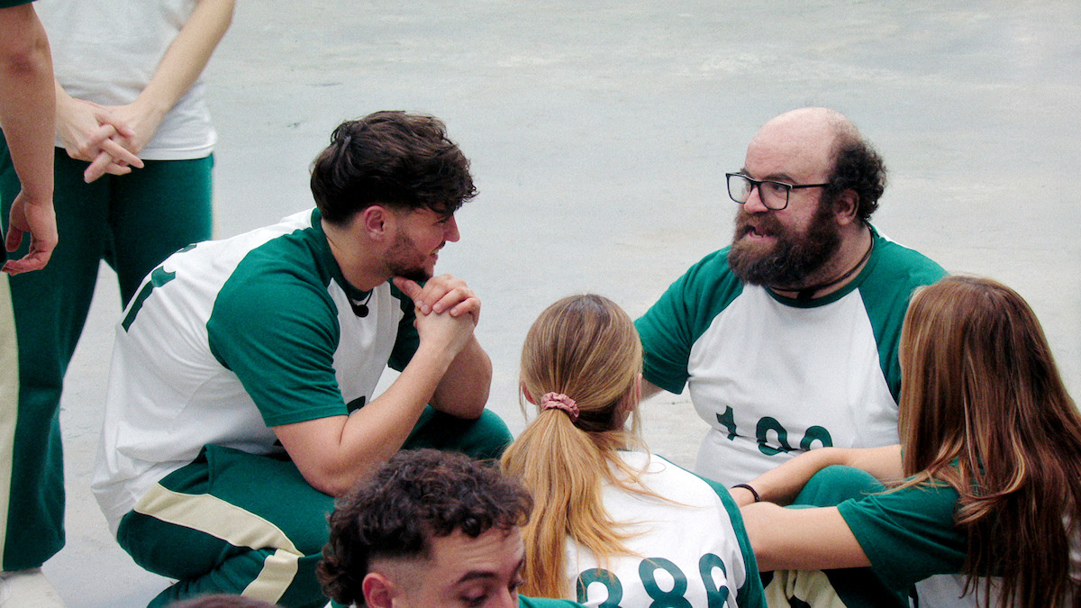 A group of people in matching green and white uniforms sit on the ground inside a bright, spacious indoor area, engaged in conversation and teamwork. The mood appears focused and collaborative.