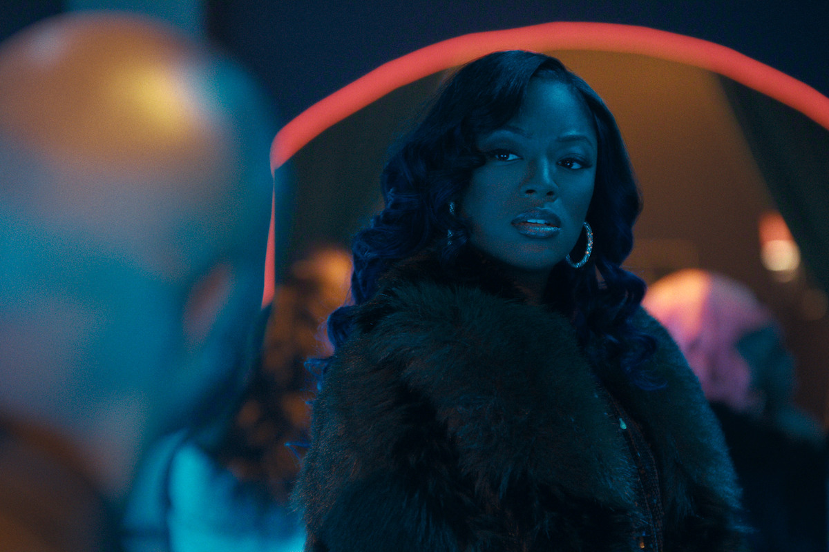 Woman with curled hair and hoop earrings in a dark fur coat stands in a dimly lit nightclub, facing a man whose head is out of focus in the foreground; vibrant blue and pink lighting creates a dramatic atmosphere.