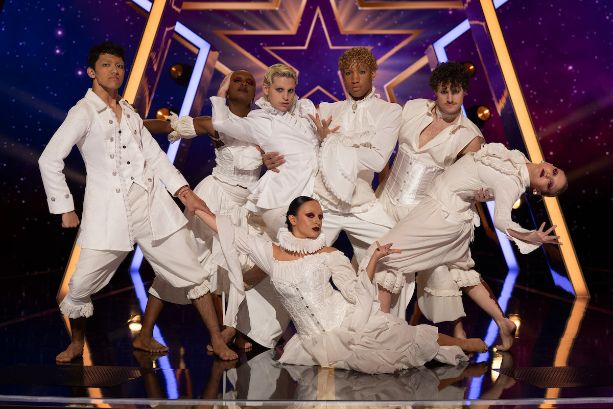 Movement 55 in dramatic white costumes pose on a shiny stage with a large star backdrop and purple lights, creating a theatrical and energetic performance atmosphere.