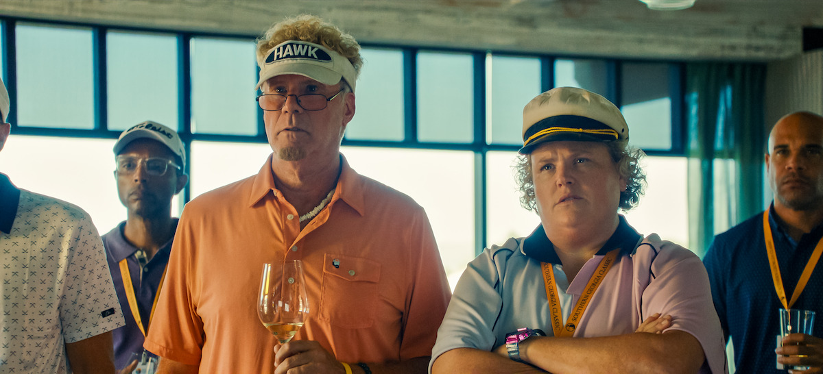 A group of people in colorful golf attire with name badges listen attentively indoors, two in front wear visors and hats, one holding a wine glass, with modern windows and sunlight in the background.